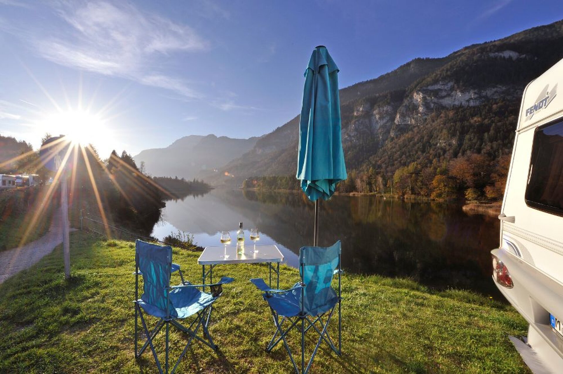 Blick auf den See mit zwei Stühlen, einem Schirm, einem Tisch und Weingläser mit Weinflasche auf der Wiese davor neben einem Wohnwagen