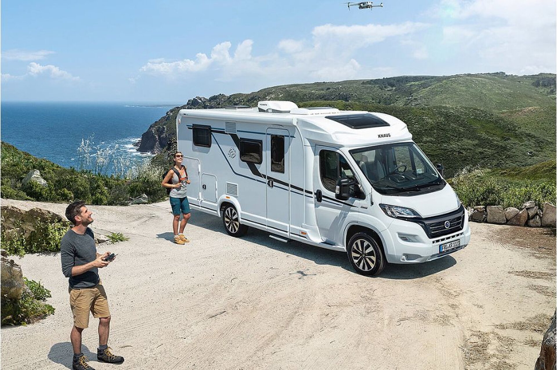 Ein Reisemobil steht an einer Küste und zwei Menschen schauen auf eine fliegende Drohne.
