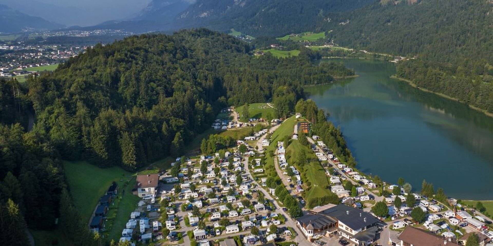Das Luftbild des Campingplatzes Seeblick Toni mit einem Waldstück, dem See und den Bergen im Hintergrund