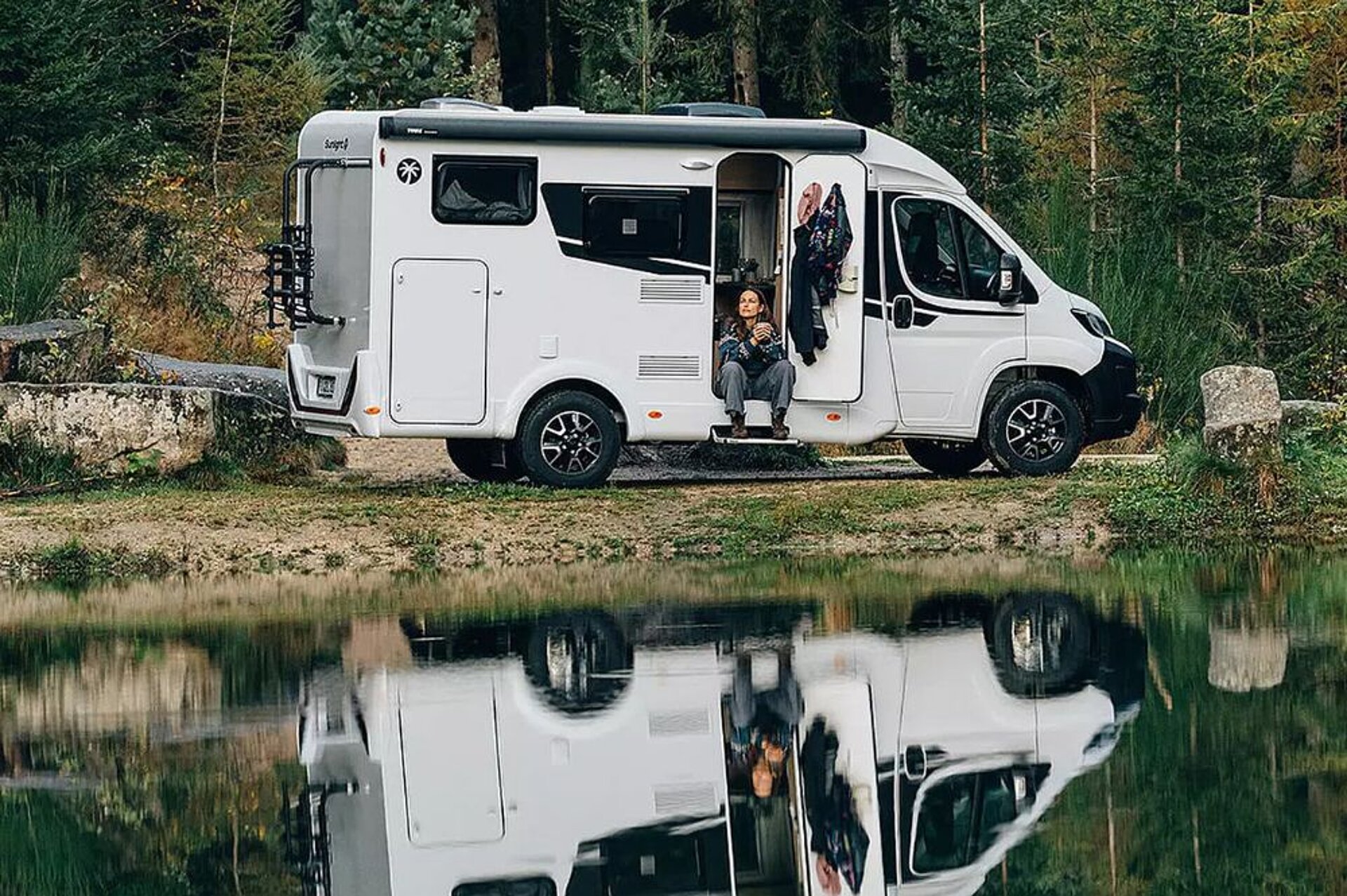 Eine Frau sitzt an der offenen Tür eines Wohnmobils welches am Wasser zwischen Steinen steht.