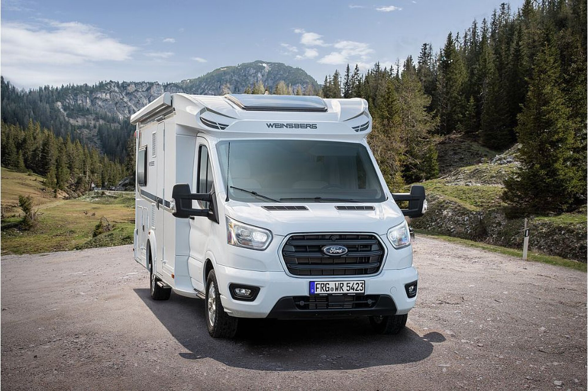 Ein Wohnmobil steht auf einem unbefestigten Platz vor Wald und Bergen