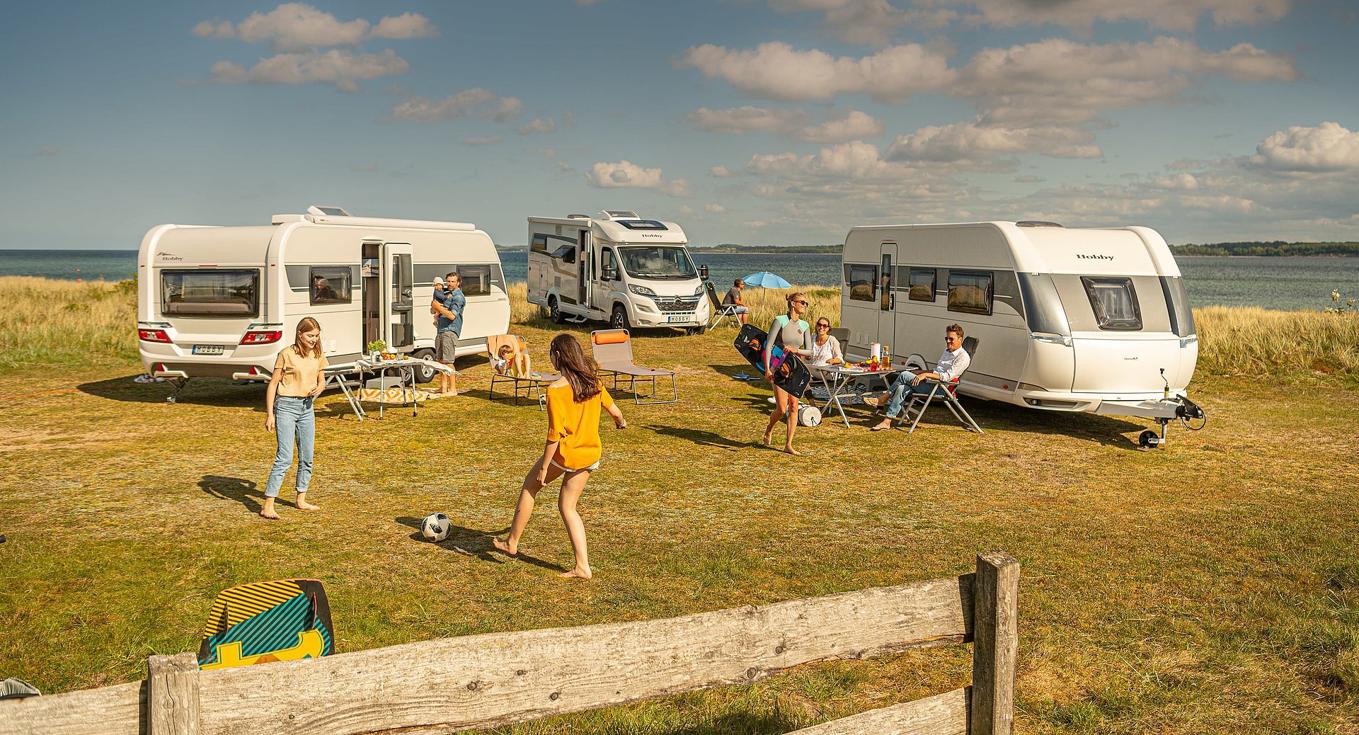 Zwei Caravane und ein Wohnmobil auf einer Wiese vor einem Meer und im Vordergrund spielen und entspannen Menschen.
