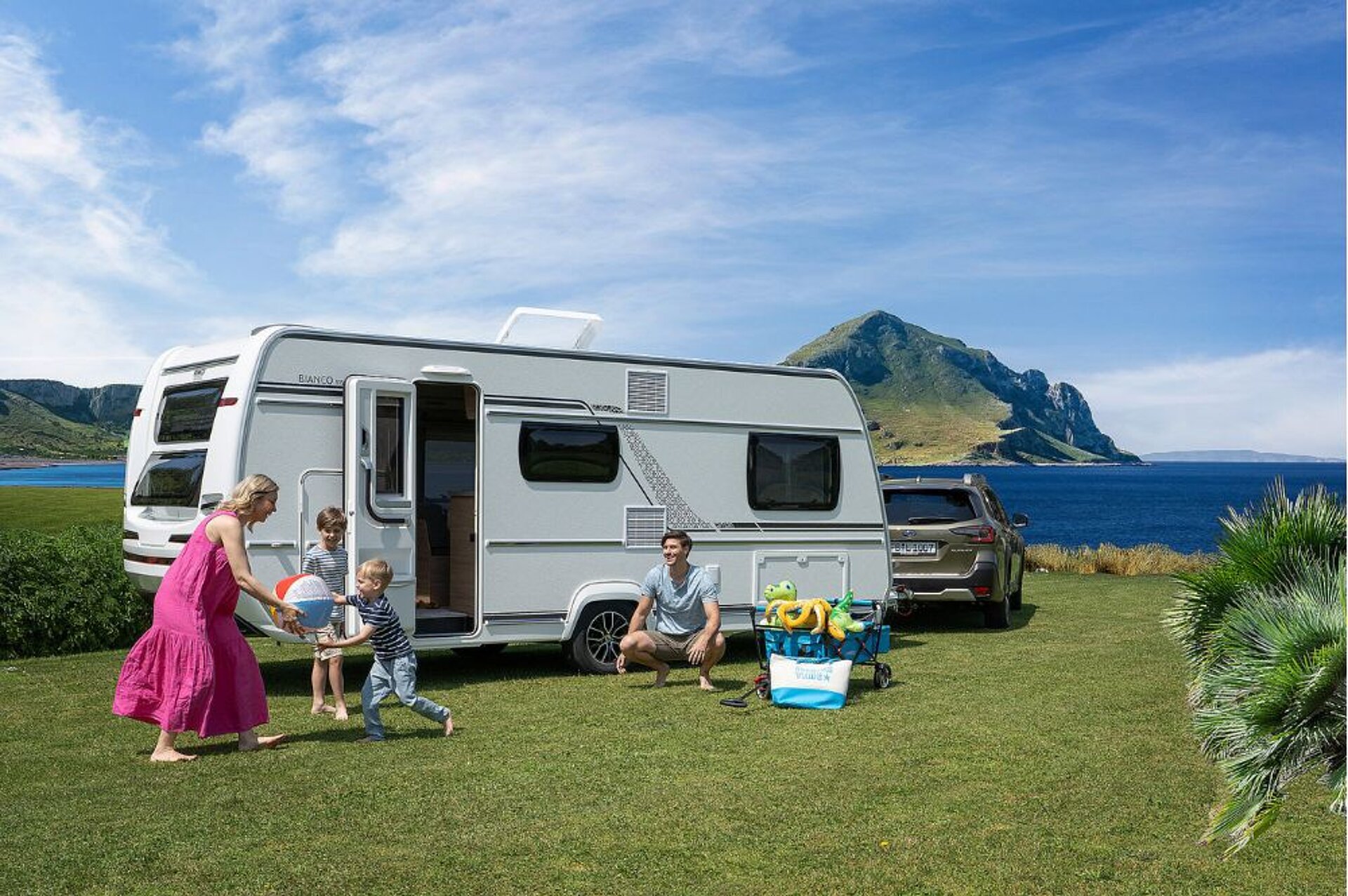 Eine Familie spielt vor einem Fendt Wohnwagen auf einer Wiese am Meer.