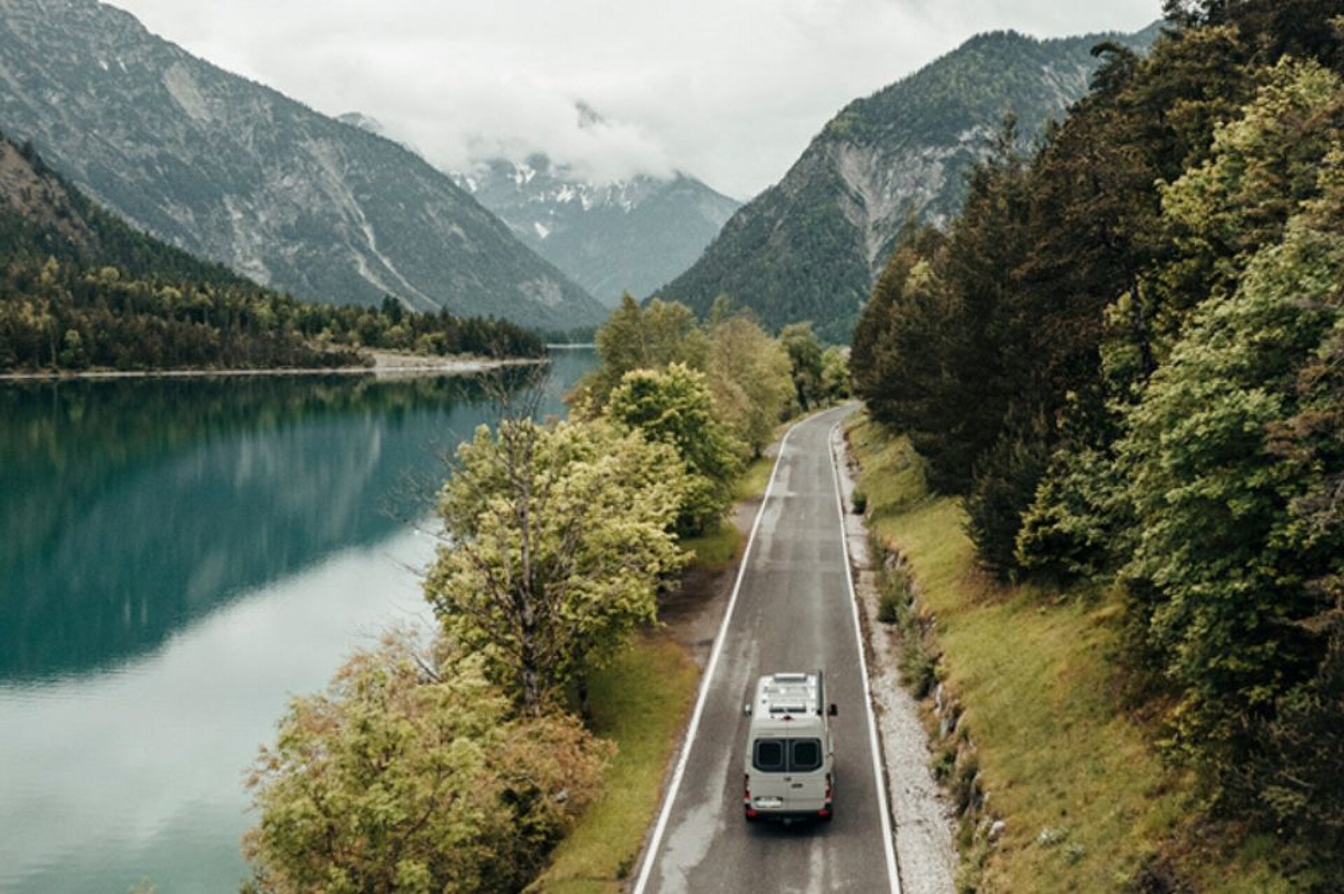 Ein Campervan fährt auf einer Straße zwischen Wasser und Wald auf die Berge zu.