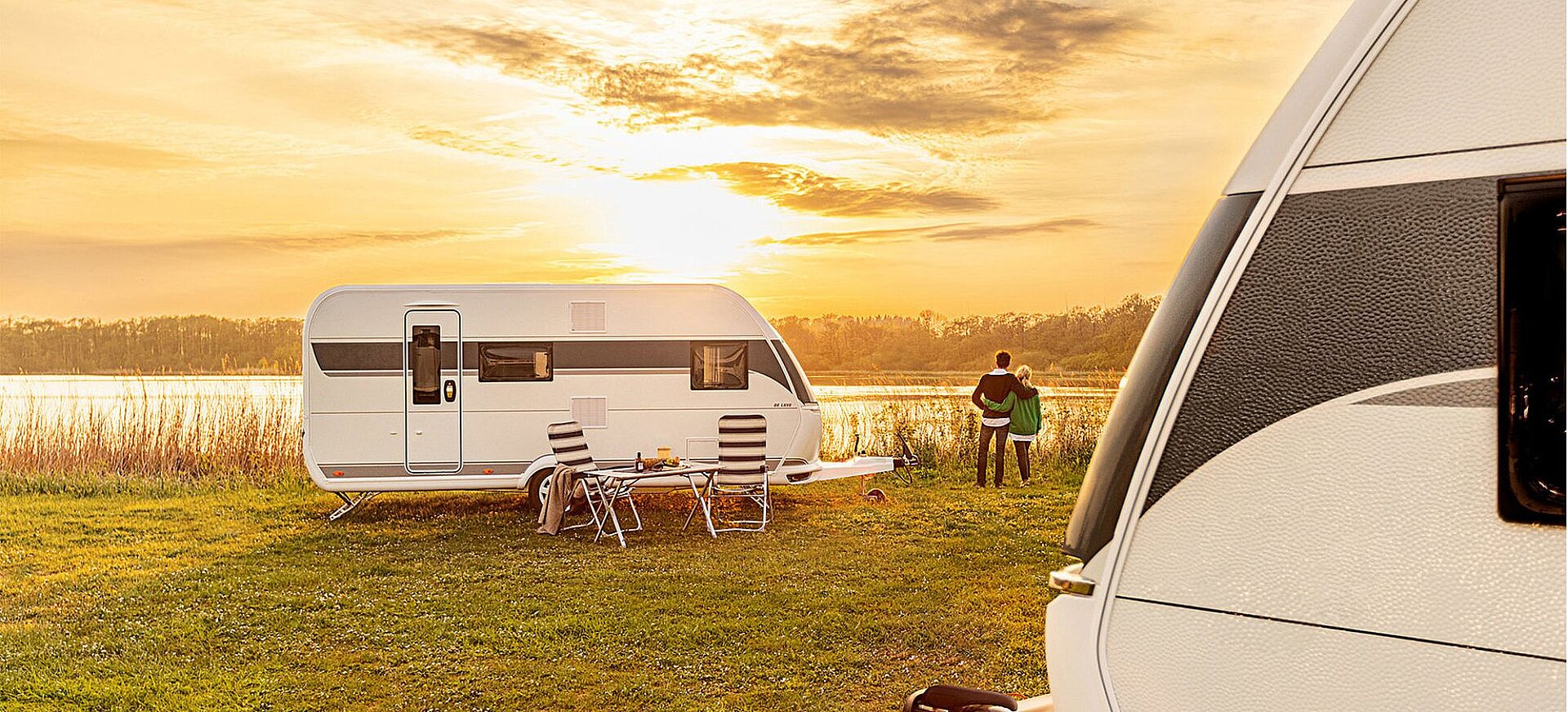 Ein Wohnwagen steht auf einer Wiese am See mit Tisch und zwei Stühlen davor