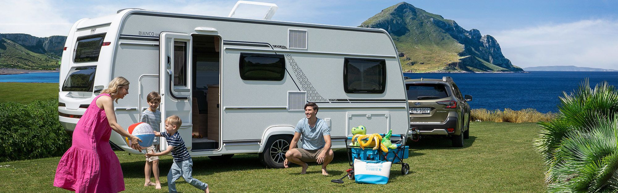 Eine Familie spielt vor einem Fendt Wohnwagen auf einer Wiese am Meer.
