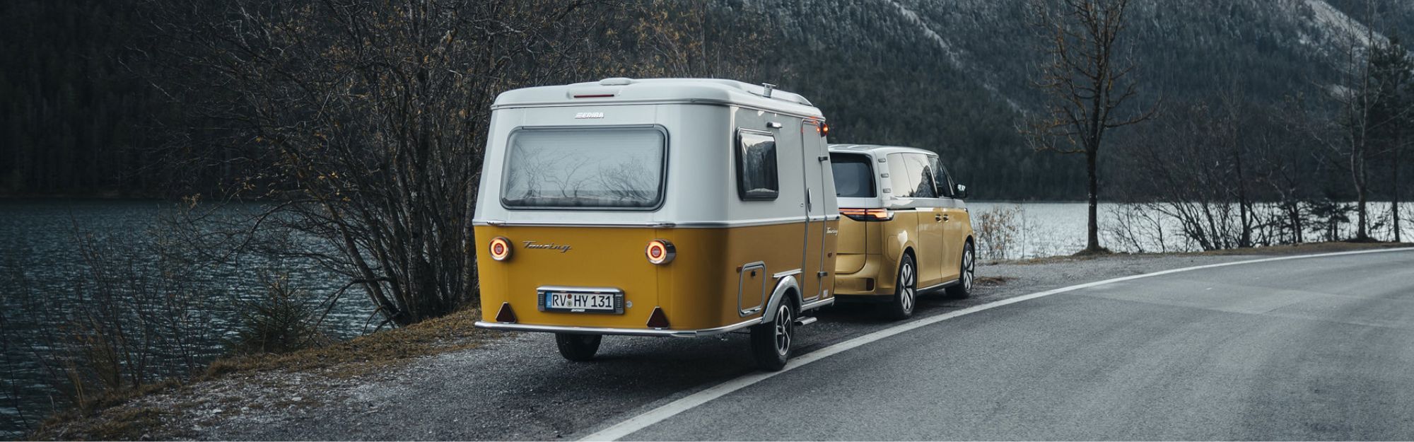 Ein Wohnwagen steht mit einem Bus vorne dran auf dem Seitenstreifen einer Straße am Wasser.
