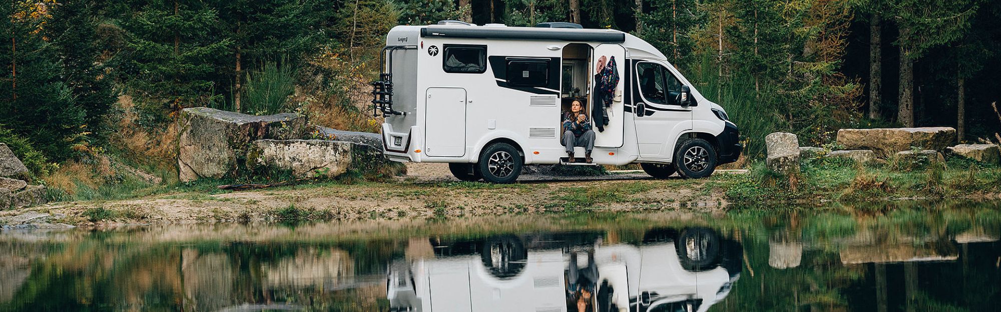 Eine Frau sitzt an der offenen Tür eines Wohnmobils welches am Wasser zwischen Steinen steht.