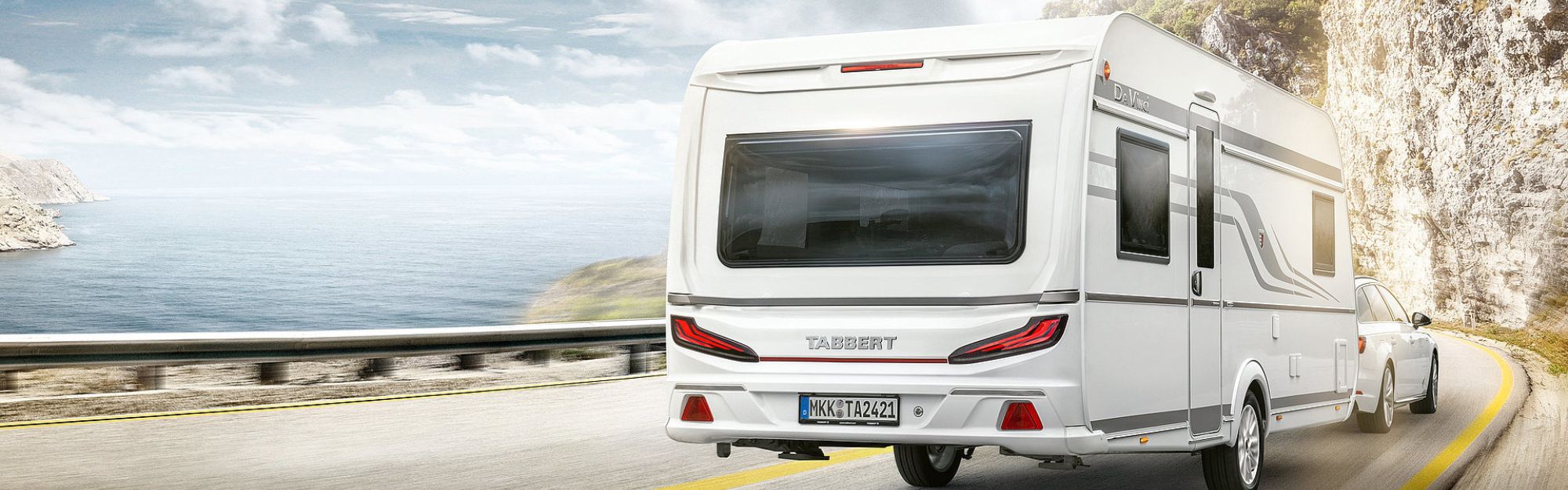 Ein Wohnwagen wird auf einer Straße zwischen Wasser und Felsen von einem Auto gezogen