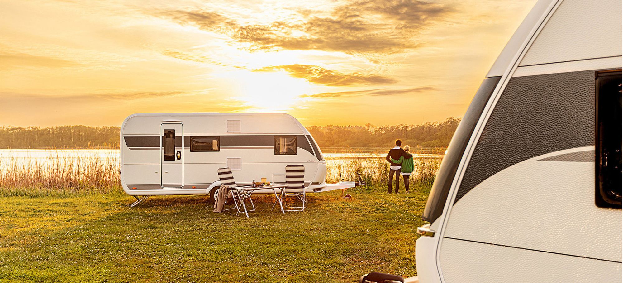 Ein Wohnwagen steht auf einer Wiese am See mit Tisch und zwei Stühlen davor