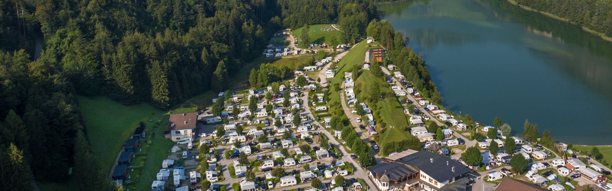 Das Luftbild des Campingplatzes Seeblick Toni mit einem Waldstück, dem See und den Bergen im Hintergrund